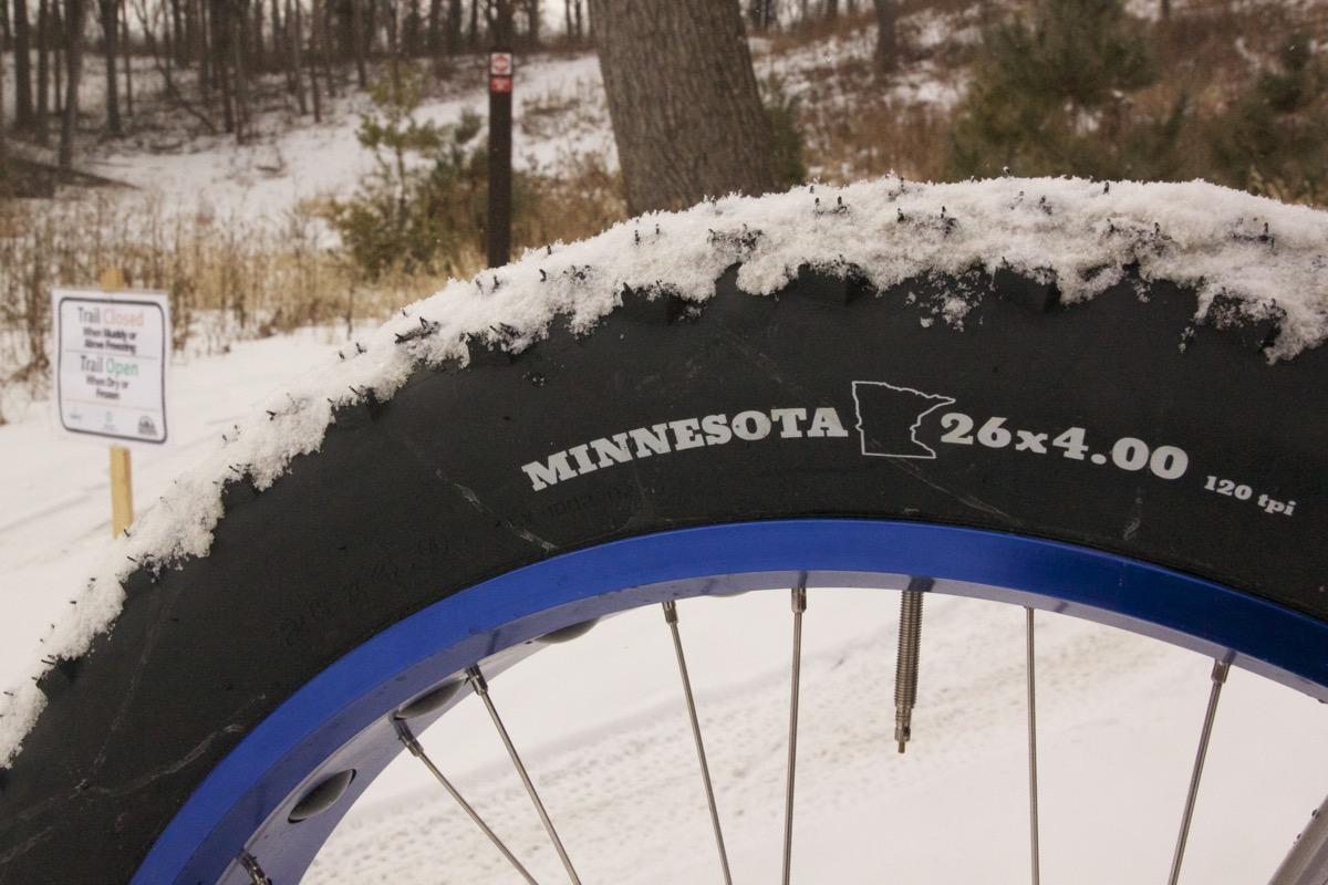 Framed Minnesota 2.0: Close-up of a fat bike tire with snow on top, featuring markings that read "MINNESOTA 26x4.00 120 tpi." In the background, a sign indicates trail conditions with "Trail Closed" and "Trail Open" instructions, surrounded by a snowy landscape.
