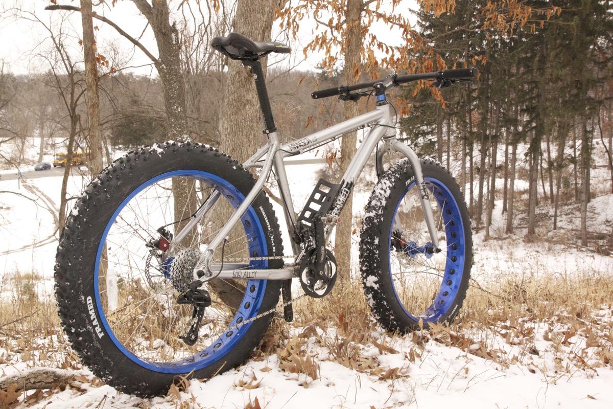 Framed Minnesota 2.0: A silver fat bike with blue rims sits in a snowy landscape, surrounded by trees and grass. The bike is partially covered in snow, showcasing its wide, studded tires designed for winter riding.