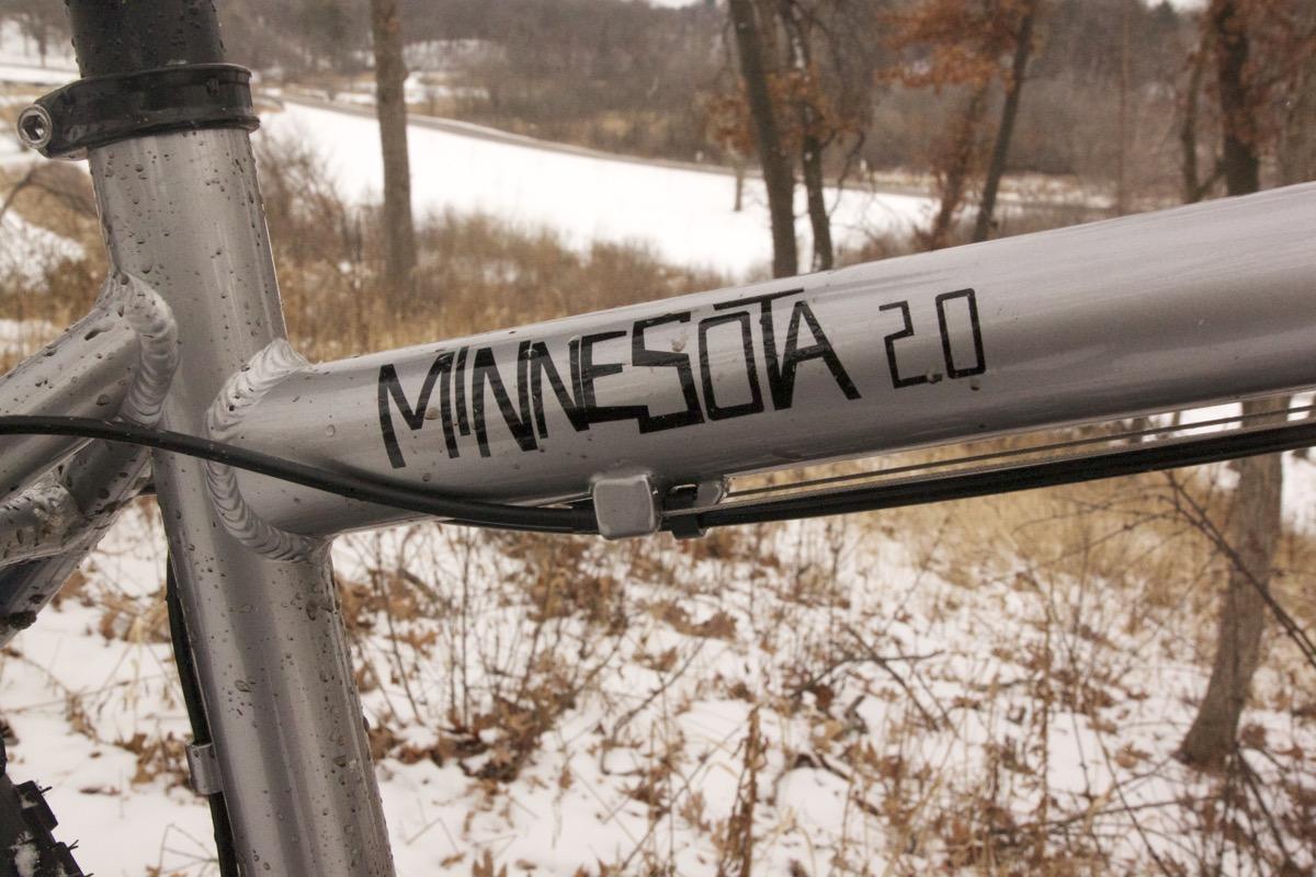 Framed Minnesota 2.0: Close-up of a bike frame featuring the text "MINNESOTA 2.0" in bold black letters. The frame has a metallic silver finish, with droplets of water on its surface, and is surrounded by a winter landscape with patches of snow and bare trees in the background.
