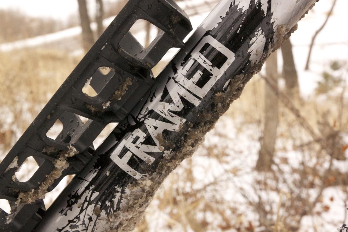 Framed Minnesota 2.0: Close-up of a black bike frame with the word "FRAMED" prominently displayed. The frame is partially covered in dirt and snow, suggesting it has been used outdoors in winter conditions. Soft, blurred natural scenery in the background includes trees and a snow-covered landscape.