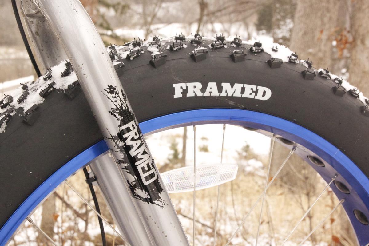 Framed Minnesota 2.0: Close-up view of a fat bike tire with winter tread, showcasing the brand "Framed" on both the tire and the bike frame. The frame has a sleek silver finish, and the tire features a blue rim. Snow is visible on the tire's surface, set against a blurred winter landscape.