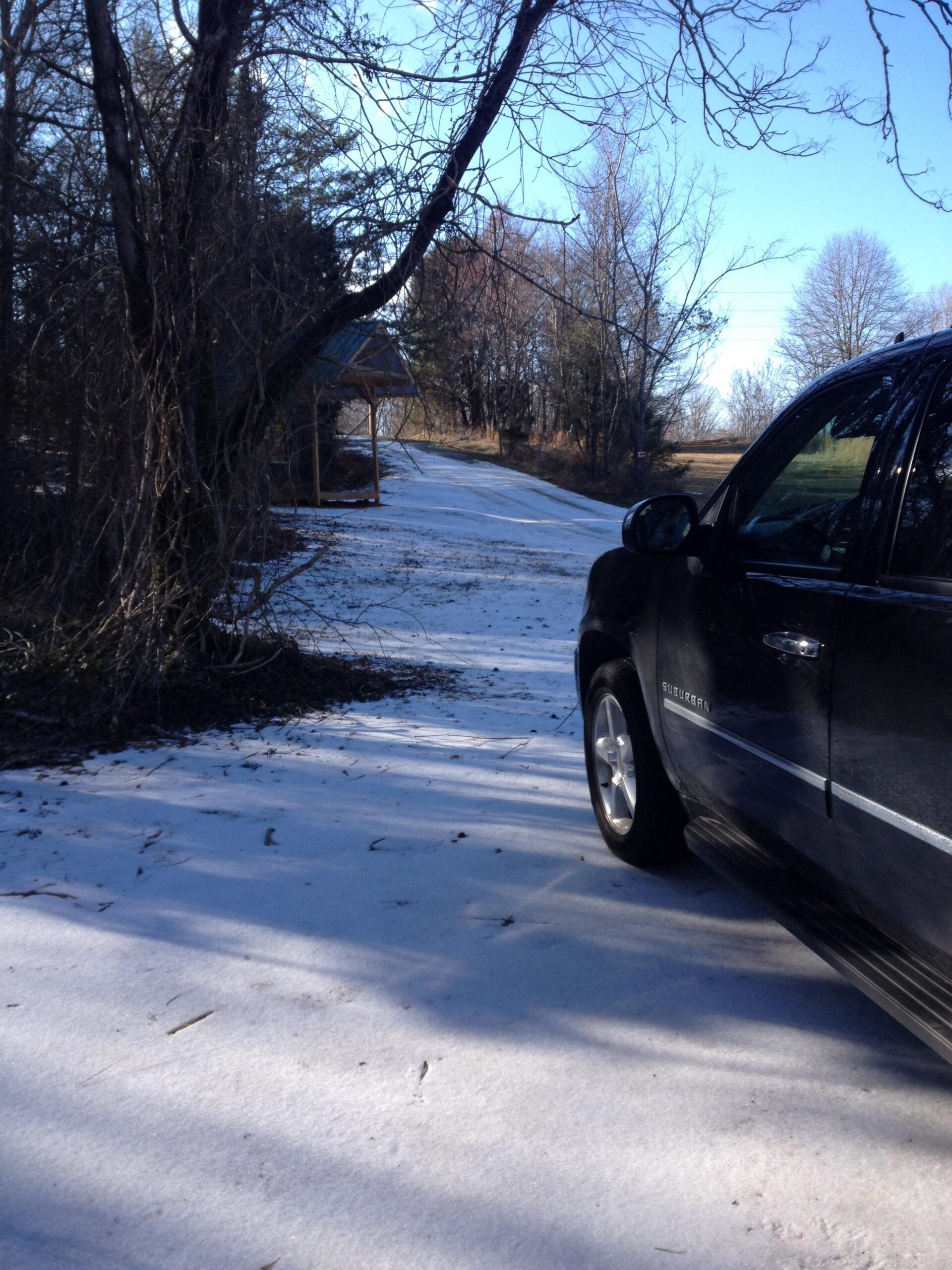 A black SUV parked on a snowy driveway with bare trees and a distant gazebo in the background under a clear blue sky. Rocky River Trail mountain bike trail.