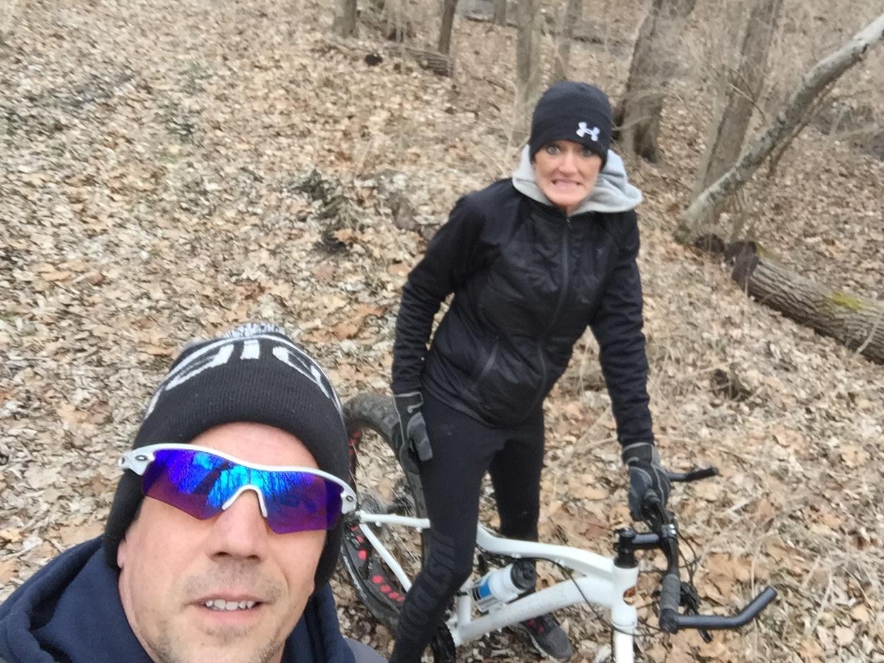 Motobecane NightTrain Bullet: Two individuals posing for a selfie in a forested area covered with fallen leaves. One person is wearing sunglasses and a black beanie, while the other is dressed in a black jacket and pants, standing next to a mountain bike. The background features trees and a wooded path, indicating an outdoor adventure.