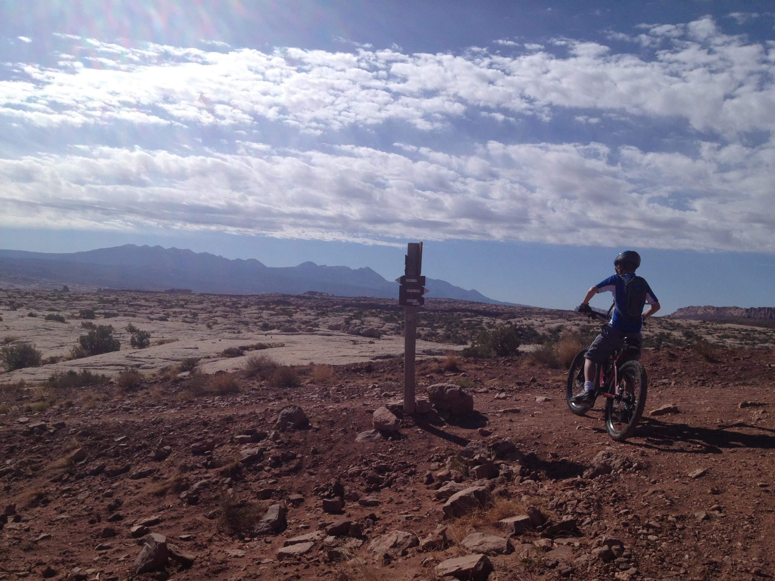 A person on a mountain bike stands next to a trail sign in a rocky, arid landscape. The background features distant mountains under a partly cloudy sky, with sunlit clouds creating a dramatic effect. The terrain is mostly dry, with sparse vegetation and scattered rocks. Moab Brand Trails mountain bike trail.