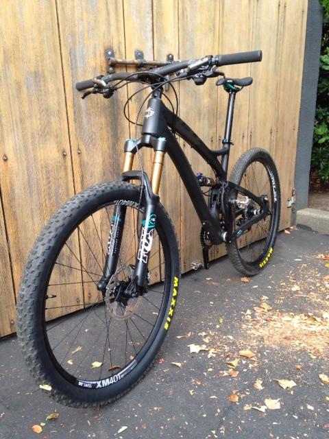 Yeti SB5c: A black mountain bike with gold front suspension forks, positioned at an angle against a wooden fence. The bike features thick tires and has various decals on the frame and wheels. There are scattered leaves on the ground around the bike, indicating an outdoor setting.