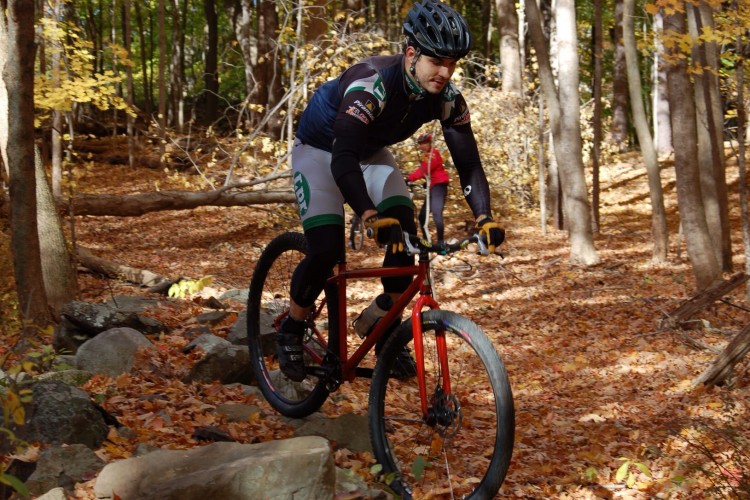 A cyclist in a black helmet rides over rocky terrain on a red mountain bike, surrounded by autumn foliage. The ground is covered in orange and yellow leaves, with trees in the background. Another rider in a red jacket is visible in the background.
