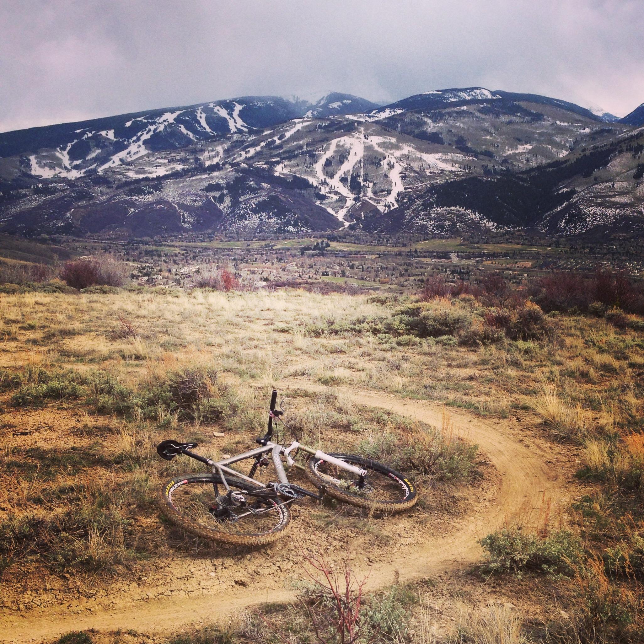 A mountain bike lies on a dirt trail surrounded by grassy terrain, with a backdrop of snow-capped mountains and a cloudy sky. The trail gently curves through the landscape, showcasing the natural beauty of the area. Berry Creek Loop mountain bike trail.