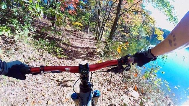 A mountain biker's perspective, riding on a narrow, leaf-covered trail alongside a clear lake, with vibrant autumn foliage in the background under a bright blue sky. Paris Mountain State Park mountain bike trail.
