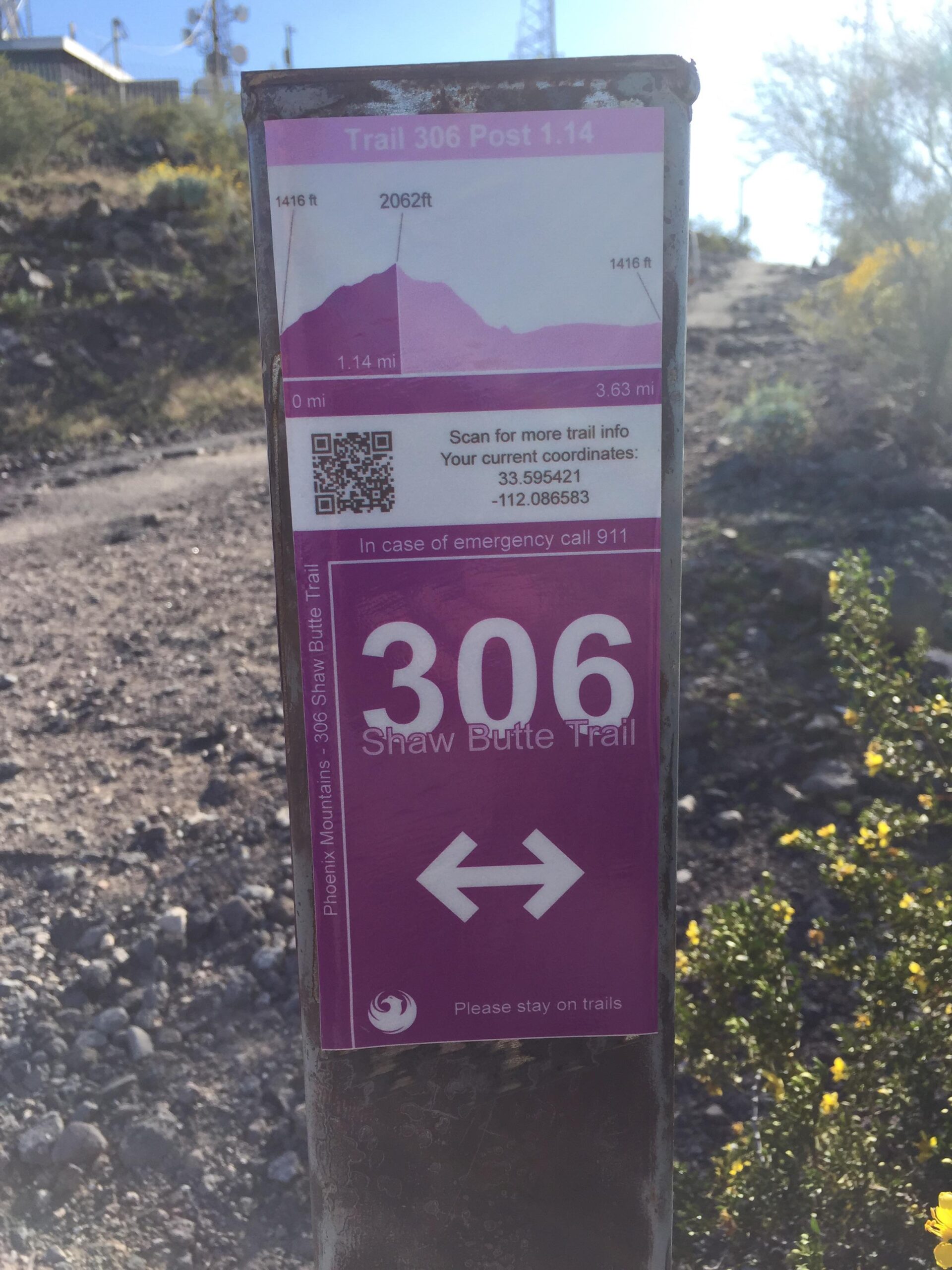 Trail marker for Shaw Butte Trail 306 at post 1.14, displaying elevation information (1416 ft to 2062 ft), trail length (1.14 mi), and QR code for additional trail information. The sign advises to stay on trails and includes emergency contact details. The background shows rocky terrain and desert vegetation under a clear blue sky. Trail #100 mountain bike trail.