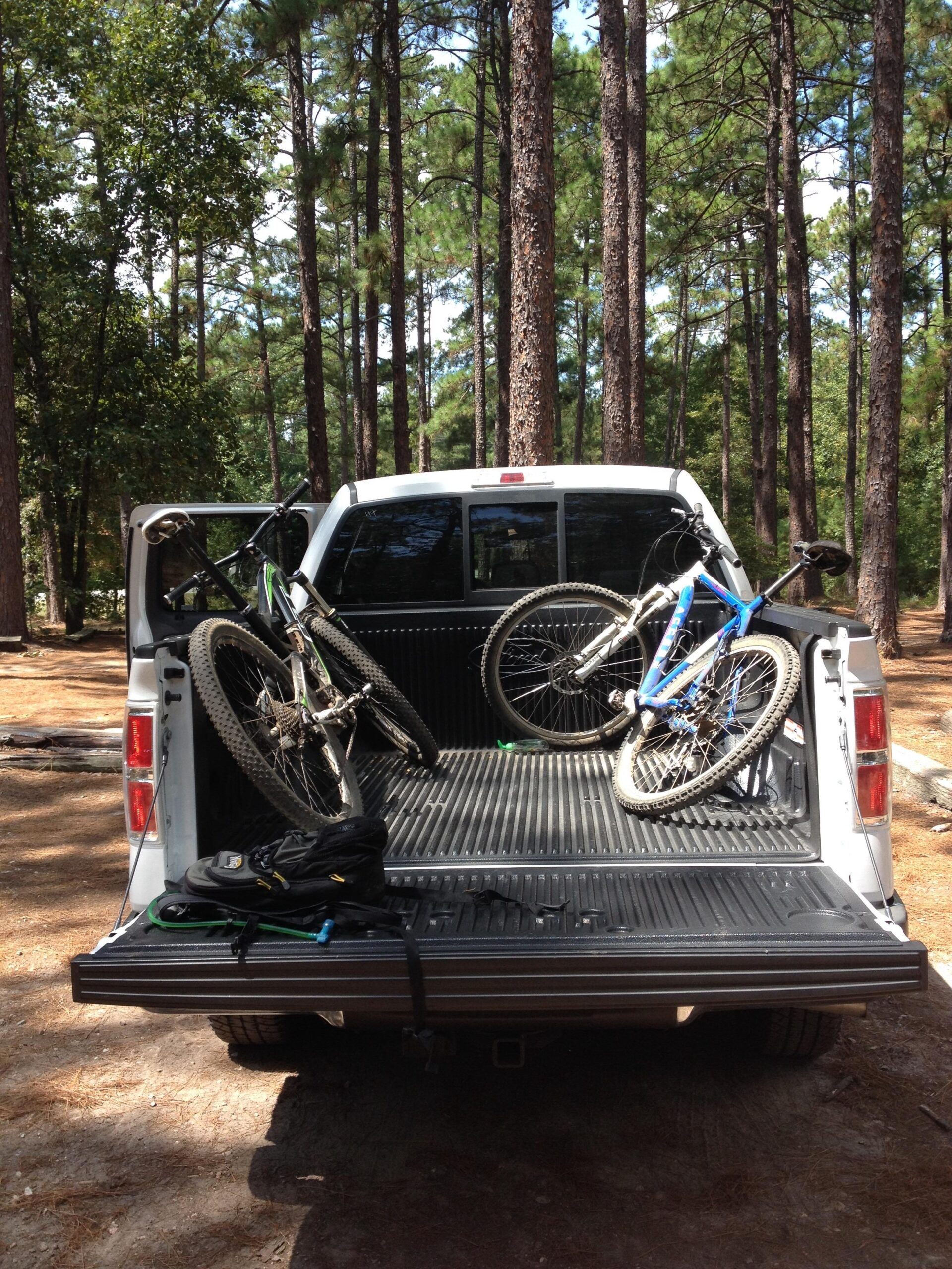 A view of the back of a white pickup truck parked in a wooded area, containing two mountain bikes secured in the truck bed, along with a black backpack resting on the edge. Tall pine trees are visible in the background under a clear sky. Harbison State Forest mountain bike trail.