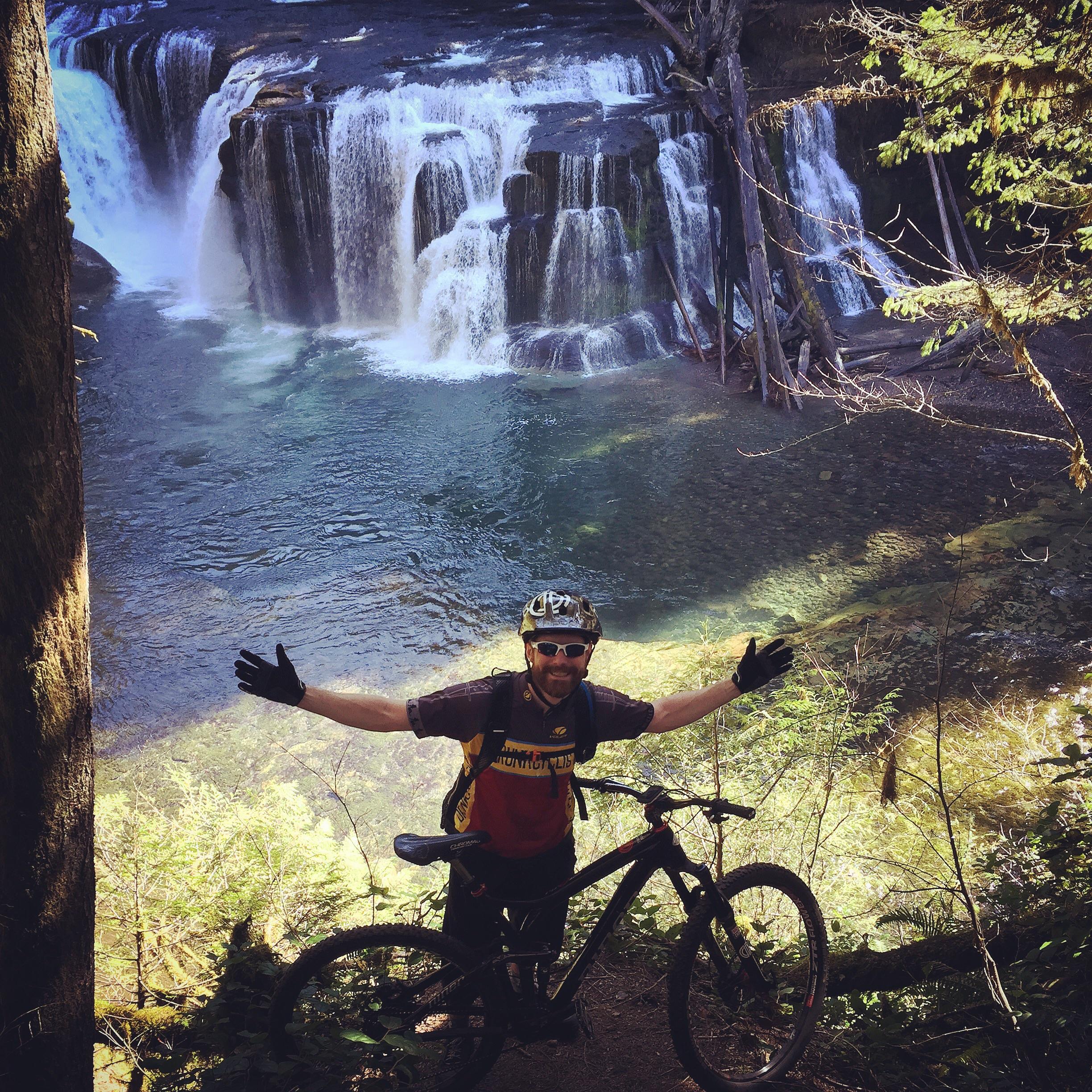 Niner R.I.P. 9 RDO: A person with a mountain bike stands with arms outstretched in front of a scenic waterfall and clear water, surrounded by lush greenery. The sunlight filters through the trees, creating a vibrant outdoor atmosphere.