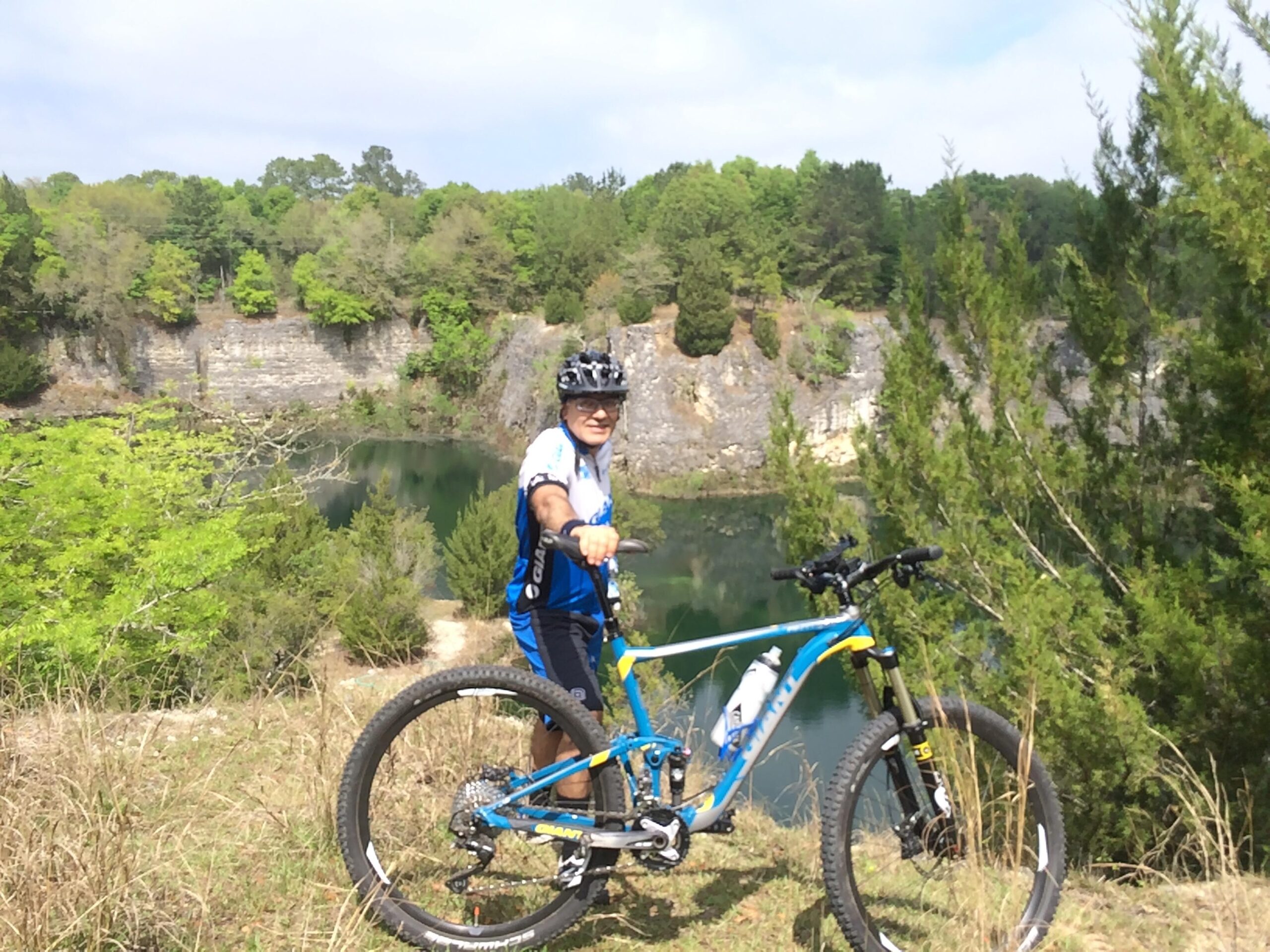 A person on a mountain bike stands on a grassy hill, smiling and posing for the camera. The background features a tranquil water body surrounded by rocky cliffs and lush green trees. Sunlight filters through the clouds, illuminating the scene. Haile's Trails mountain bike trail.