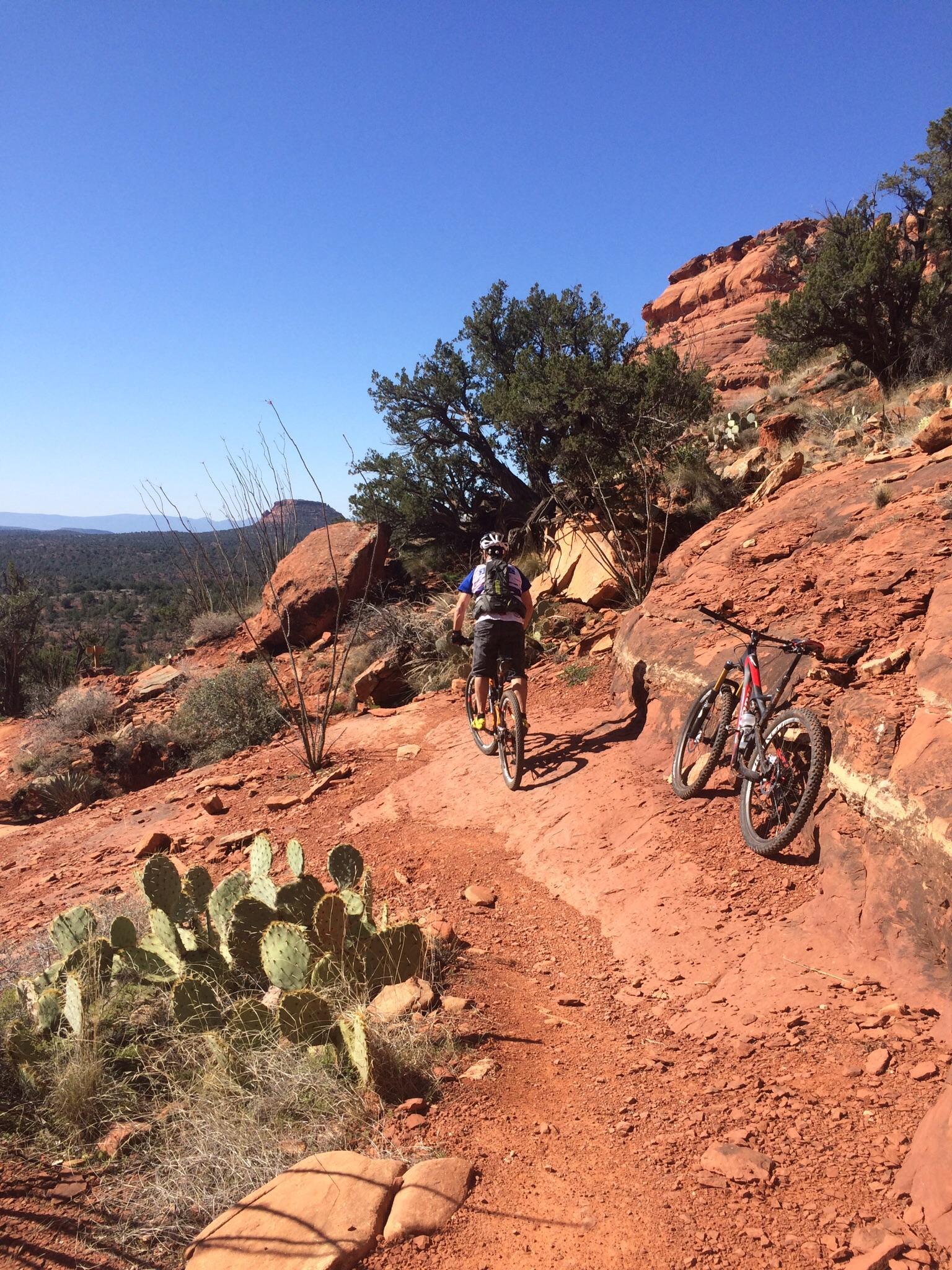 A mountain biker riding along a rocky trail in a desert landscape, with red rock formations and a clear blue sky in the background. Two mountain bikes are parked on the trail, alongside a cactus. Mescal Trail mountain bike trail.
