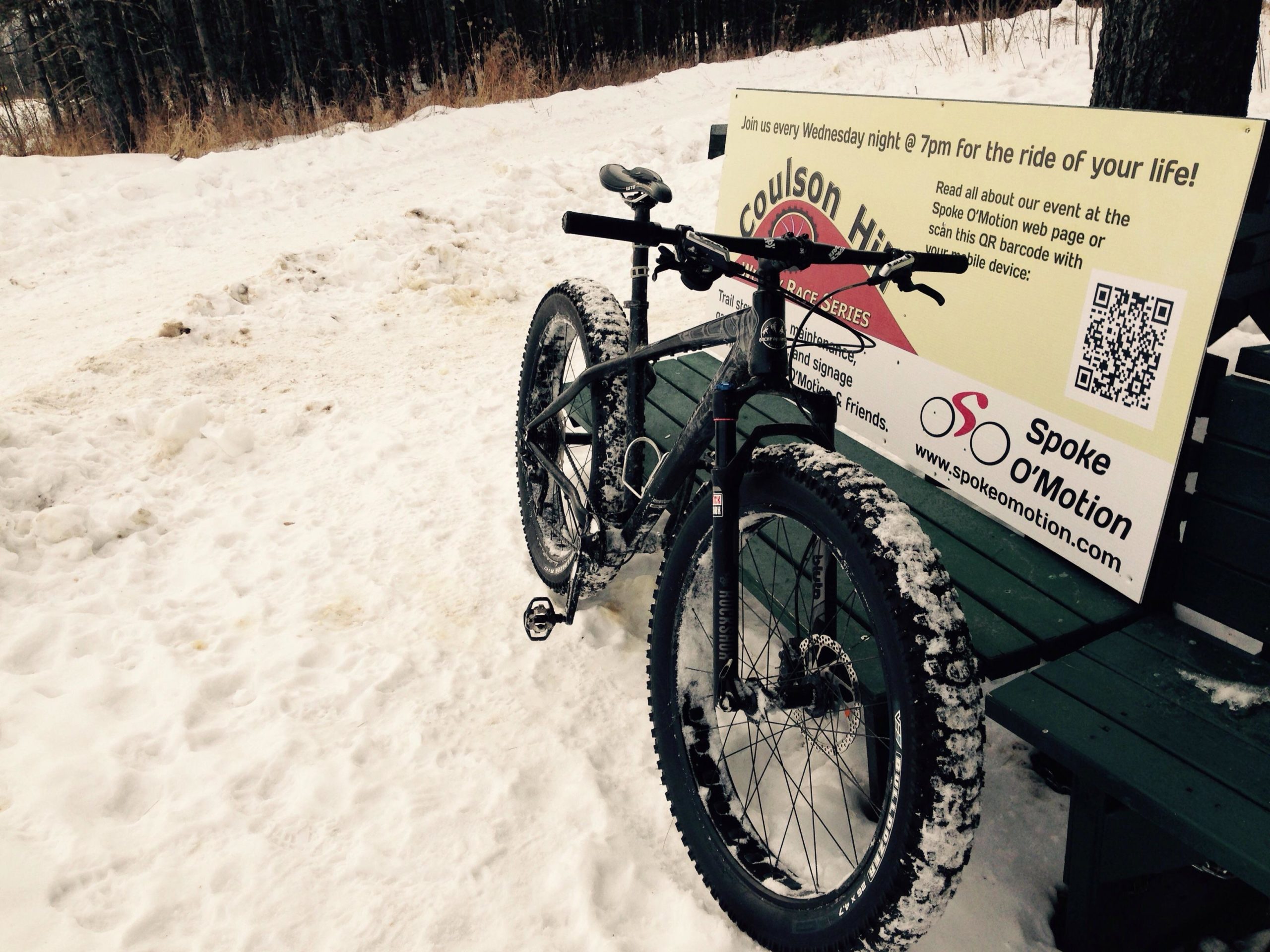 A fat bike with snow-covered tires is parked beside a large sign promoting weekly bike rides at Coulson Hill, with details about the event and a QR code for more information. The background features a snowy landscape and trees, indicating a winter setting. Coulson's Hill mountain bike trail.