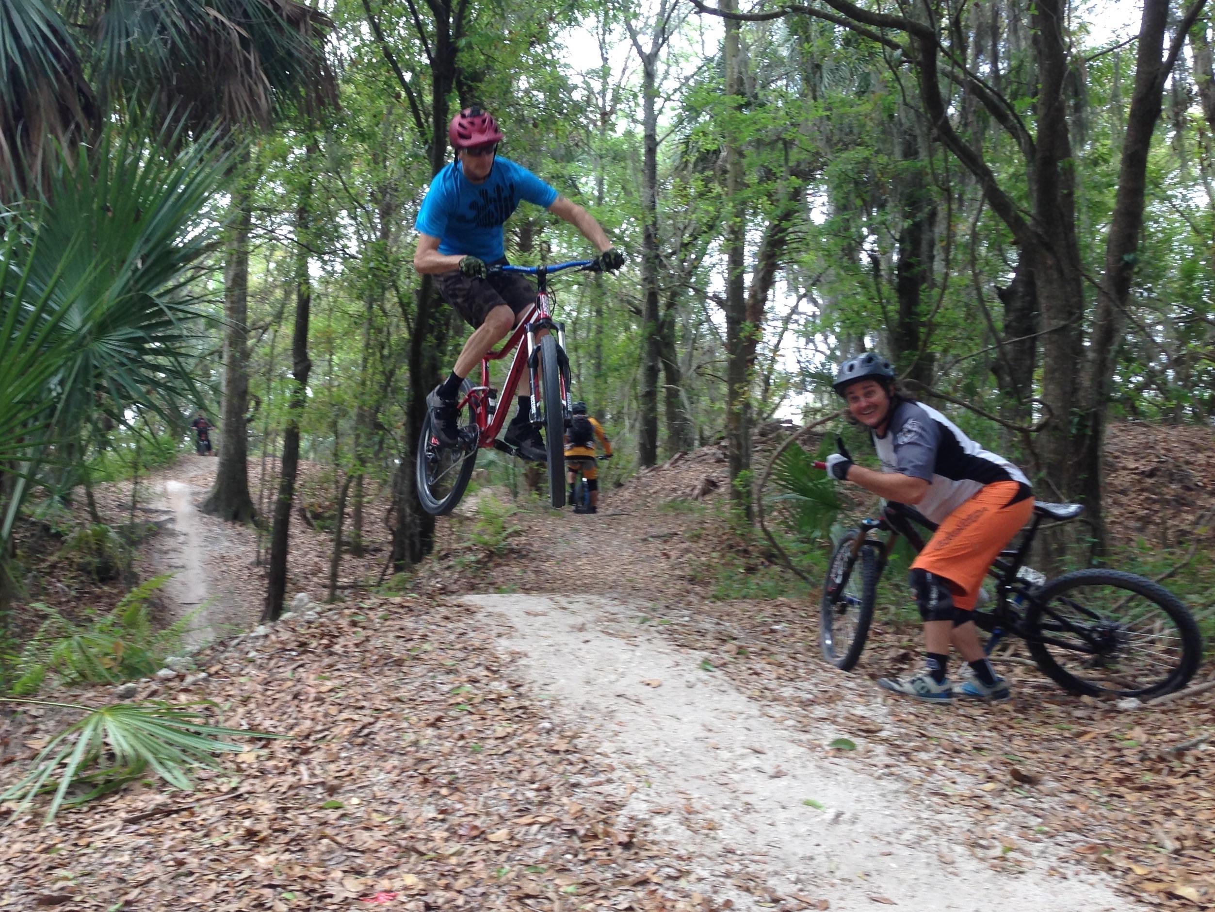 Two mountain bikers in a forested trail. One rider in blue is mid-air, jumping off a small ramp on a red bike, while a second rider in an orange and black outfit smiles and gives a thumbs-up while standing beside their bike. The background features trees and a winding dirt path. Alafia River State Park mountain bike trail.