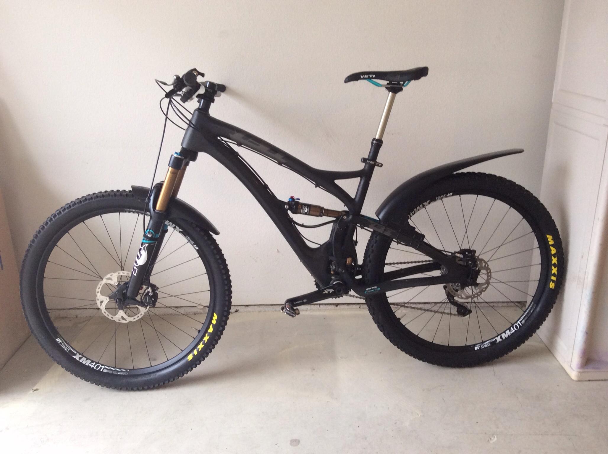 Yeti SB5c: A black mountain bike with a sleek frame and prominent front suspension, featuring thick tires and a disc brake system. The bike is positioned against a light-colored wall, showcasing its modern design and components, including a raised seat and a front fender.