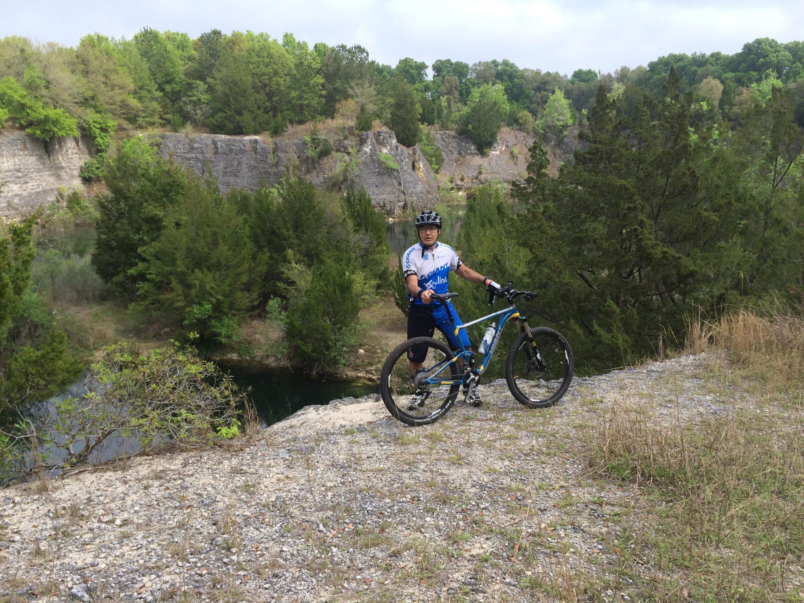 A person in cycling gear stands next to a mountain bike on a rocky ledge overlooking a body of water, surrounded by trees and a cliff in the background. The scene captures a natural landscape with greenery and a cloudy sky. Haile's Trails mountain bike trail.