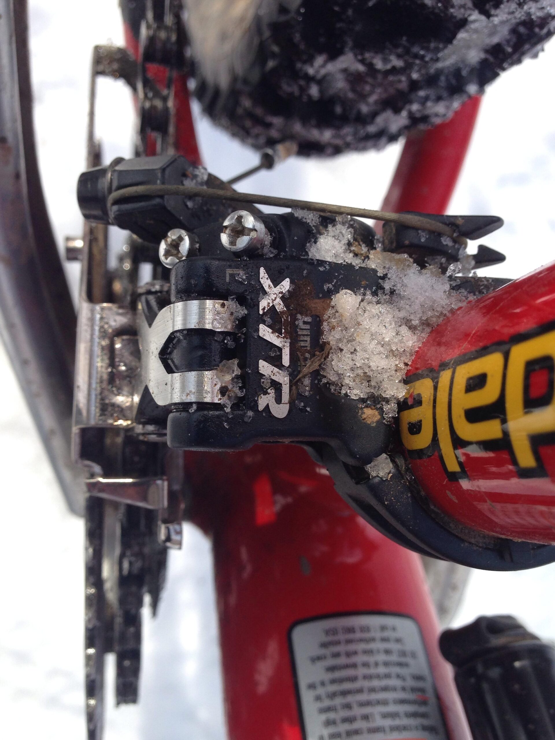 Cannondale F700: Close-up view of a bike's rear derailleur and chainring, covered in snow and ice, with red bike frame visible in the background. The derailleur is labeled "XTR," indicating a high-performance component, and there are metal screws and detailed parts showing wear from use in winter conditions.