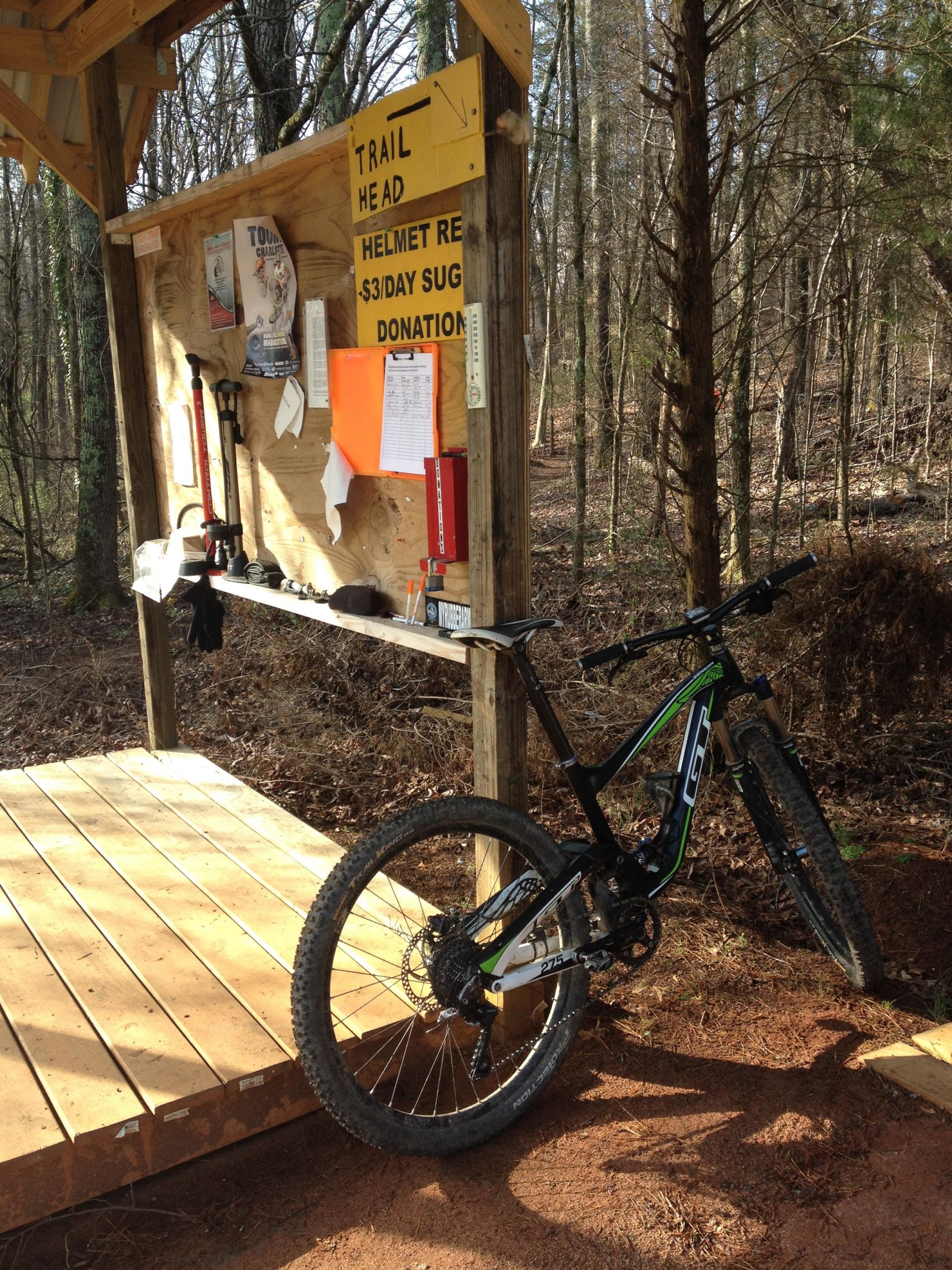 A mountain bike parked beside a wooden trailhead sign, which includes information on helmet rental and suggested donations. The background features a wooded area with trees and underbrush, and the ground is covered in dirt and leaves. Rocky River Trail mountain bike trail.
