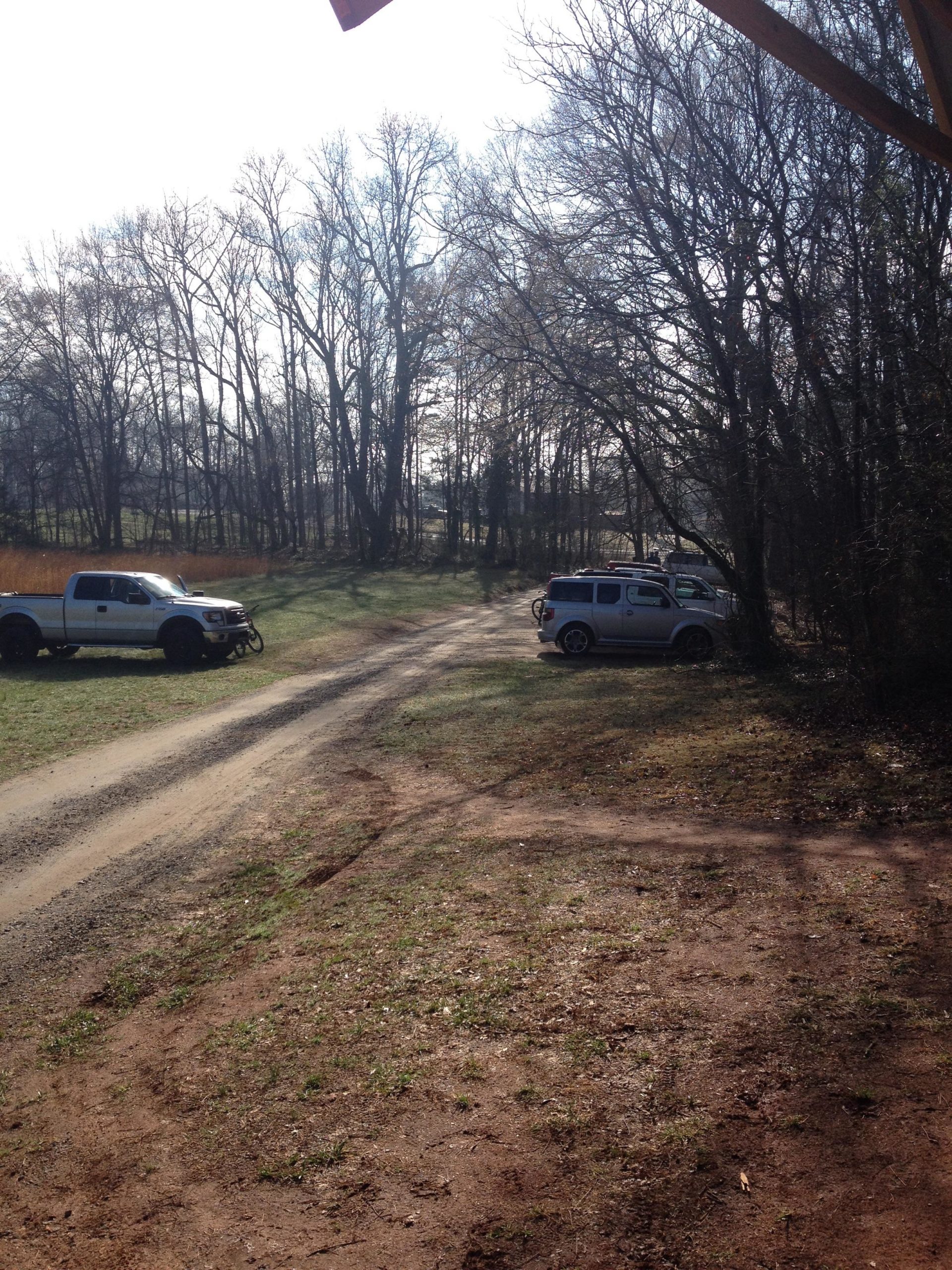 A dirt path leads through a wooded area, with two vehicles parked along the side. On the left, a white pickup truck is visible, while a silver SUV is parked on the right. The background features bare trees and overcast skies, creating a serene, rural atmosphere. Rocky River Trail mountain bike trail.