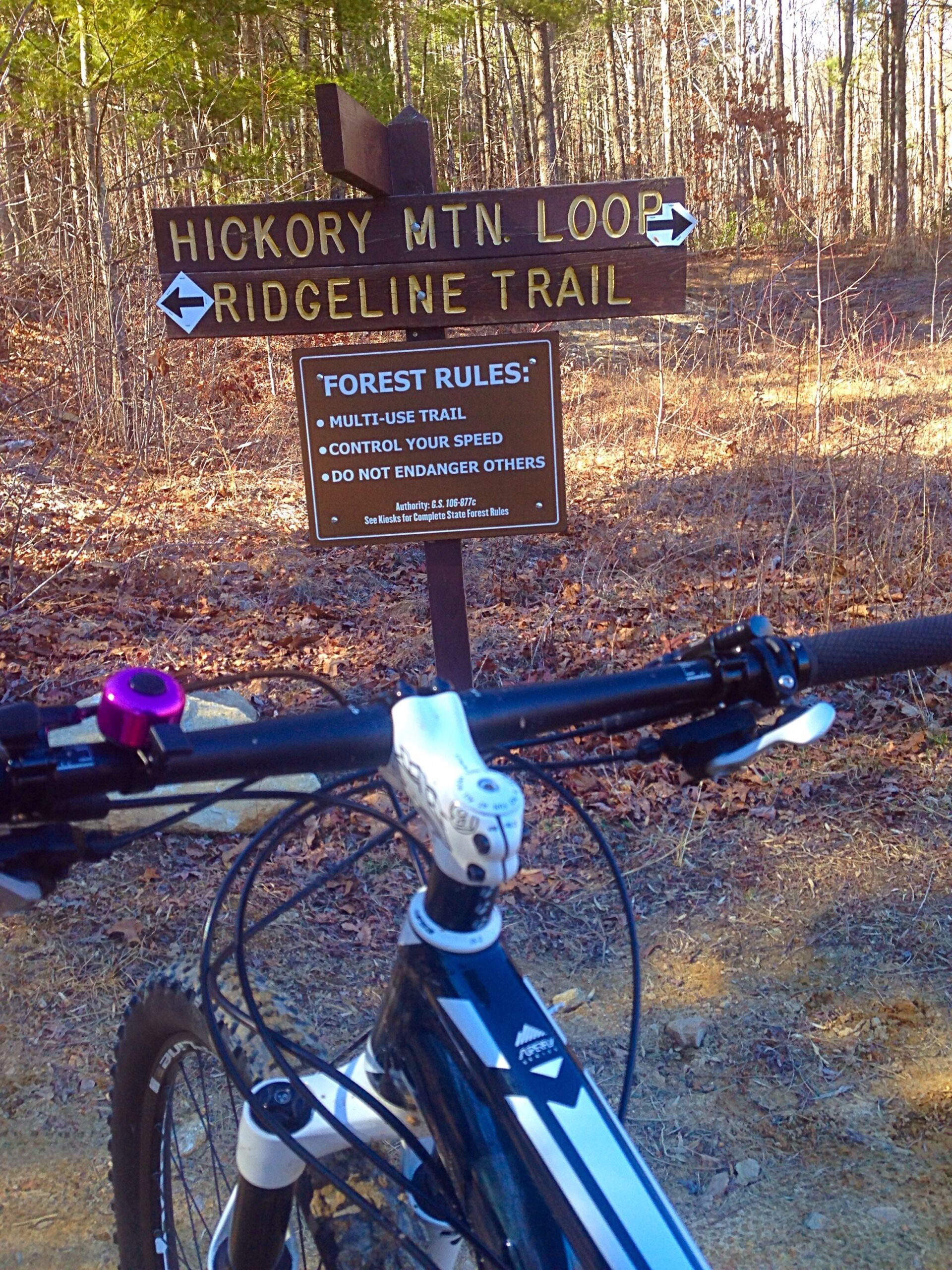 Image of a mountain bike parked at a trail intersection sign indicating "Hickory Mtn. Loop" to the right and "Ridgeline Trail" to the left. Additional signage outlines forest rules including "MULTI-USE TRAIL," "CONTROL YOUR SPEED," and "DO NOT ENDANGER OTHERS," with a wooded background and fallen leaves on the ground. Hickory Mountain Loop mountain bike trail.