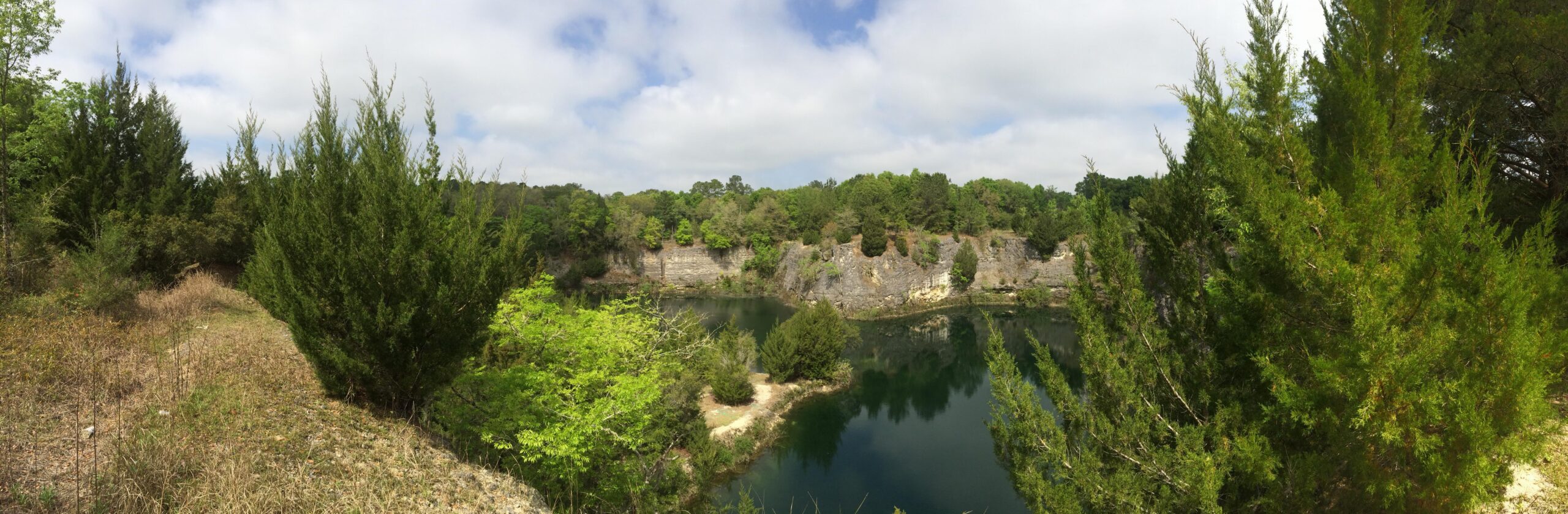 A panoramic view of a tranquil lake surrounded by green trees and rocky cliffs under a partly cloudy sky. The scene depicts a natural landscape with a mix of lush foliage and exposed rock formations, reflecting a serene and peaceful environment. Haile's Trails mountain bike trail.