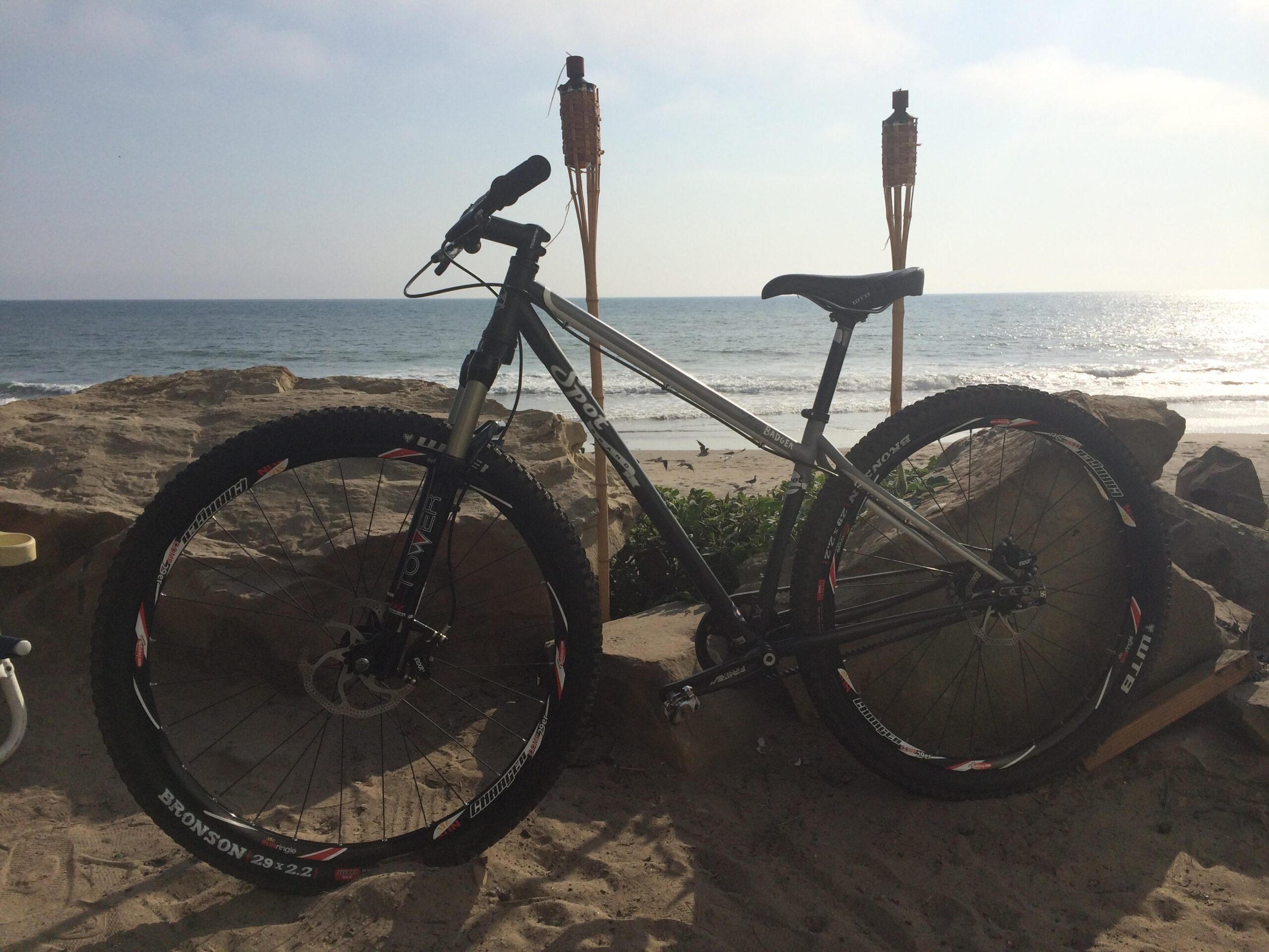 SPOT Honey Badger Singlespeed: A mountain bike resting on a rocky outcrop near the beach, with the ocean and a sandy shoreline visible in the background. Two tiki torches are positioned nearby, and the scene is illuminated by soft, natural light.
