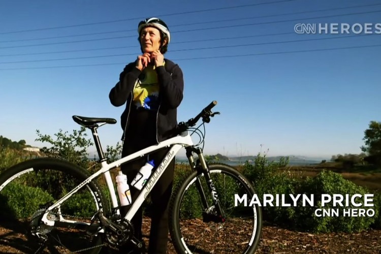 A woman wearing a helmet and casual clothing stands beside a white mountain bike in a scenic outdoor setting, ready to ride. The background features lush greenery and a clear blue sky. Text on the image identifies her as a CNN Hero.