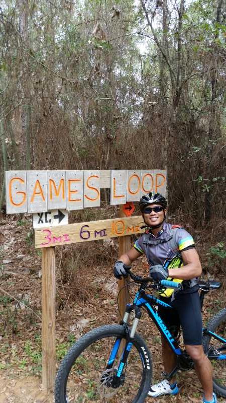 A cyclist in a colorful outfit stands next to a wooden sign marked "GAMES LOOP," indicating various trail lengths of 3 miles, 6 miles, and 10 miles. The background features a dense forest with trees and underbrush. The cyclist is wearing a helmet and sunglasses, smiling while holding onto their mountain bike. Games Loop mountain bike trail.