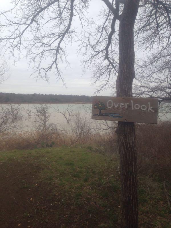 A wooden sign labeled "Overlook" is attached to a tree, overlooking a calm body of water. The scene is set in a natural area with sparse trees and a cloudy sky, indicating a serene outdoor environment. Cedar Hill State Park At Joe Pool Lake mountain bike trail.