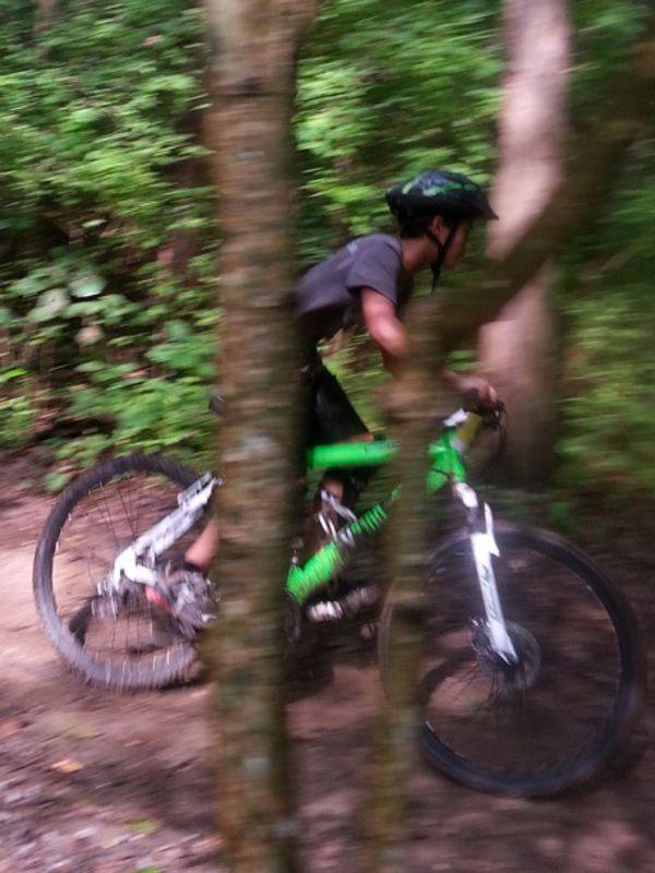 A blurred image of a young cyclist navigating a dirt trail through a forest. The bike is primarily green with white accents, and the background features dense greenery, indicating a natural setting. Tree trunks partially obscure the view, adding to the sense of movement and speed. Lock 4 mountain bike trail.