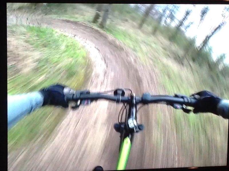 A close-up view of mountain bike handlebars as the rider navigates a winding dirt trail in a forested area. The surrounding landscape shows blurred greenery, indicating speed and motion. The image captures the exhilaration of biking through nature. Wessington Warrior Trail mountain bike trail.