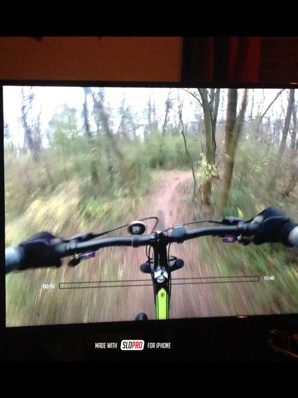 Alt text: A close-up view of bicycle handlebars on a mountain biking trail, displayed on a television screen. The scene shows a forested area with a dirt path winding through the trees. A timer is visible at the bottom of the screen. Wessington Warrior Trail mountain bike trail.