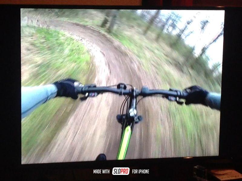 A dynamic view from a mountain bike as it navigates a winding trail through a forest. The image captures the handlebars and part of the rider's arms in motion, emphasizing speed and movement on a dirt path surrounded by trees. Wessington Warrior Trail mountain bike trail.
