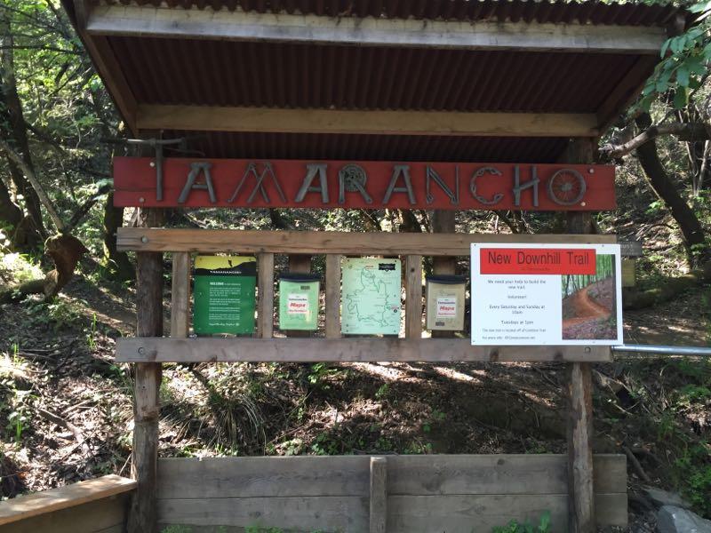 Sign for Tamarancho trailhead, featuring a prominently displayed red sign with the name "TAMARANCHO." Below the sign are informational boards with maps and trail details, alongside a notice for a new downhill trail. The background consists of trees and natural greenery typical of a forested area. Camp Tamarancho mountain bike trail.