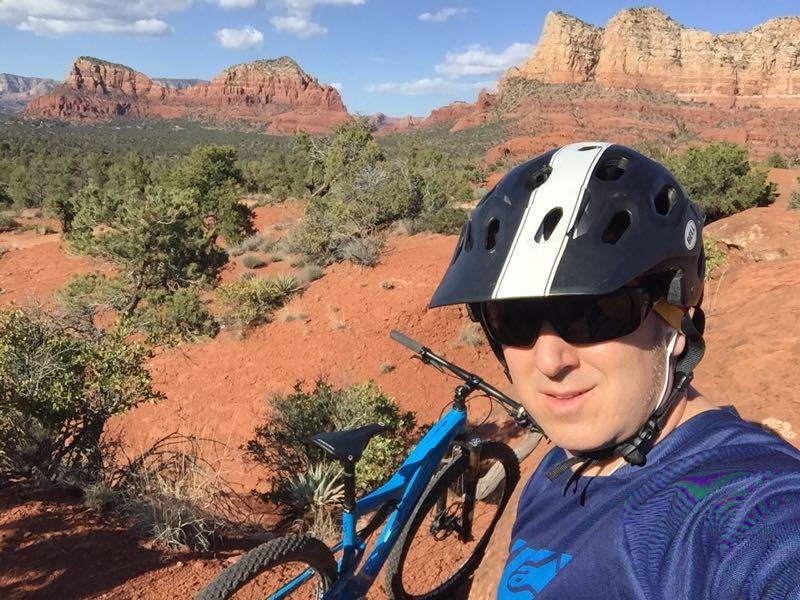 A person wearing a black helmet and a blue shirt stands next to a mountain bike in a desert landscape. The background features red rock formations and vegetation under a clear blue sky with a few clouds. Bell Rock Area Trails mountain bike trail.