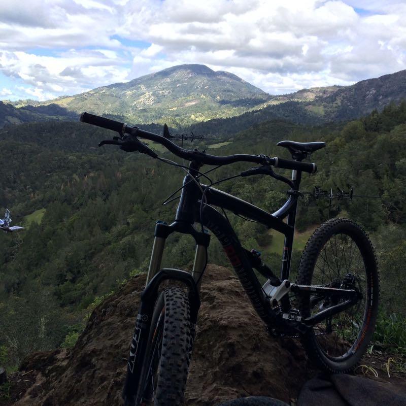 A mountain bike resting on a rock outcrop, with a scenic view of mountains and forested hills in the background under a partly cloudy sky. Oat Hill Road mountain bike trail.