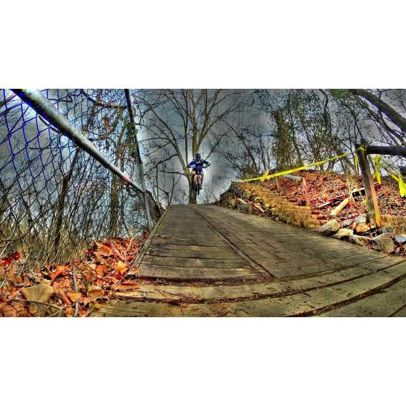 A mountain biker in a blue outfit rides down a steep wooden ramp surrounded by trees and autumn leaves. A chain-link fence runs alongside the trail, and caution tape is visible on the side, indicating a designated biking area. The sky is overcast, adding to the dramatic atmosphere of the scene. Buttermilk mountain bike trail.