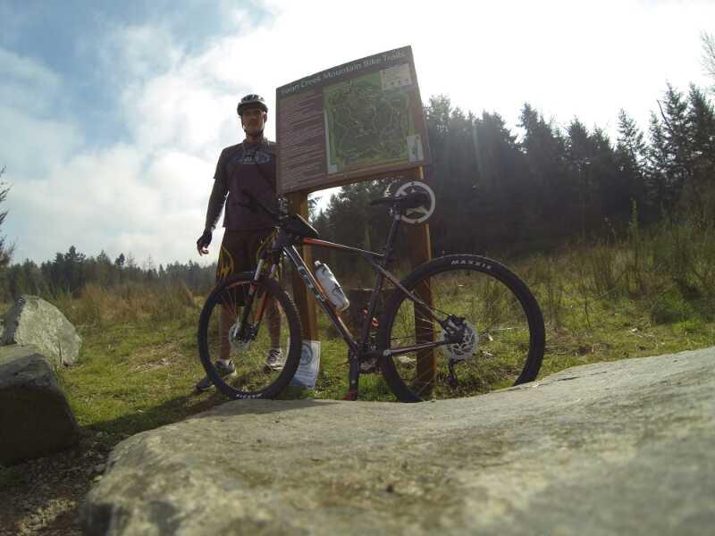 A cyclist stands next to a mountain bike and a trail map sign in a wooded area. The cyclist is wearing a helmet and sports gear, while the bike features a water bottle and mountain bike tires. Sunlight filters through the trees, and the scenery is grassy with a rocky foreground. Swan Creek mountain bike trail.