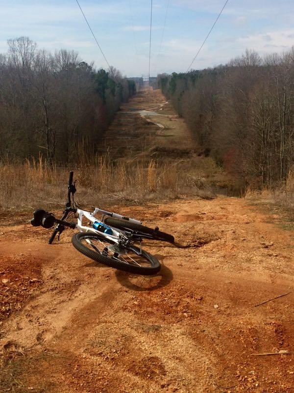 A mountain bike is leaned against the ground on a dirt slope, overlooking a wide, open clearing that stretches into the distance. Power lines run overhead, and the landscape is framed by bare trees and sparse underbrush, suggesting a rural area during late fall or winter. The color palette features earthy tones of brown and muted greens, with a cloudy blue sky above. Harbins Park mountain bike trail.