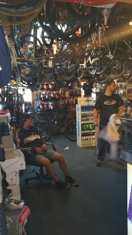 A busy bike shop interior filled with various bicycles hanging from the ceiling and displayed on the walls. Two men are present: one is sitting in a chair, looking relaxed, while the other is standing and interacting with items at the counter. Shelves stocked with bike accessories and gear are visible in the background, creating a vibrant atmosphere.