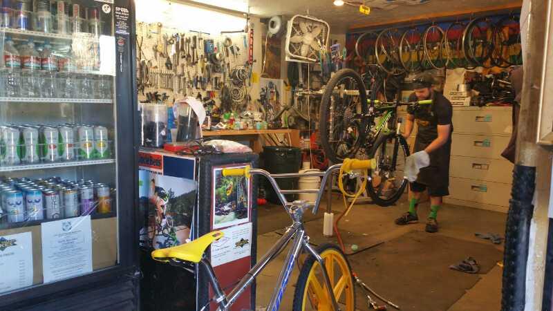 A bicycle repair shop interior featuring a variety of tools hanging on the wall and several bicycles. A man is working on a bike, holding a cloth and wearing a helmet. In the foreground, there is a brightly colored bicycle with a yellow seat. A refrigerator stocked with drinks is visible on the left side of the image.