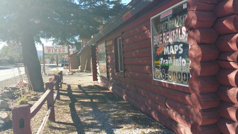 A log cabin-style building with signs advertising bike rentals and maps. The scene is sunny, with trees nearby and a dirt path alongside the structure. A wooden fence runs along the front, providing a rustic outdoor setting.