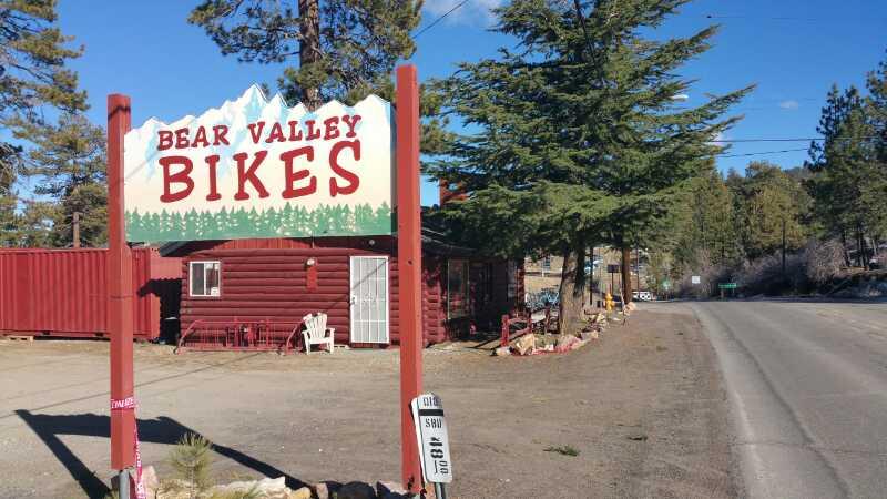 **Alt text:** A sign reading "Bear Valley Bikes" stands in front of a rustic, red wooden building surrounded by trees. The scene is set on a clear day with blue skies and a paved road nearby.