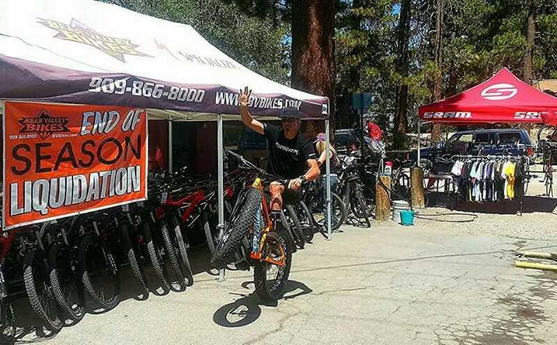 A cyclist performing a wheelie on a mountain bike in front of a tent displaying "End of Season Liquidation" at a bike shop. Various bicycles are lined up for sale in the background, and apparel is displayed on racks nearby under a sunny sky with trees in the vicinity.