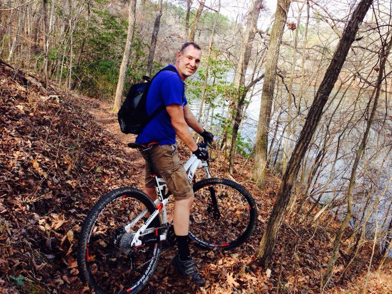 A person riding a mountain bike on a wooded trail beside a lake, wearing a blue shirt, shorts, and a backpack. The background features trees and fallen leaves, creating a natural outdoor setting. Swayback Bridge Trail mountain bike trail.