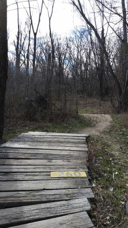 A wooden footbridge leads into a wooded area, marked with the number "260." The surrounding landscape features bare trees and a winding dirt path that disappears into the distance. The sky is partially cloudy, adding a serene atmosphere to the scene. Lawrence Riverfront Trails mountain bike trail.