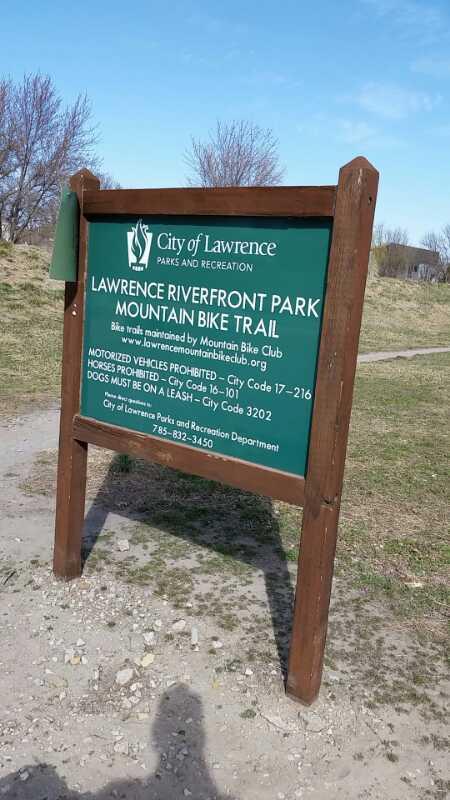 Sign at Lawrence Riverfront Park Mountain Bike Trail, detailing park regulations such as a prohibition on motor vehicles and horses, and stating that dogs must be leashed. The sign features the City of Lawrence Parks and Recreation logo and contact information. Lawrence Riverfront Trails mountain bike trail.