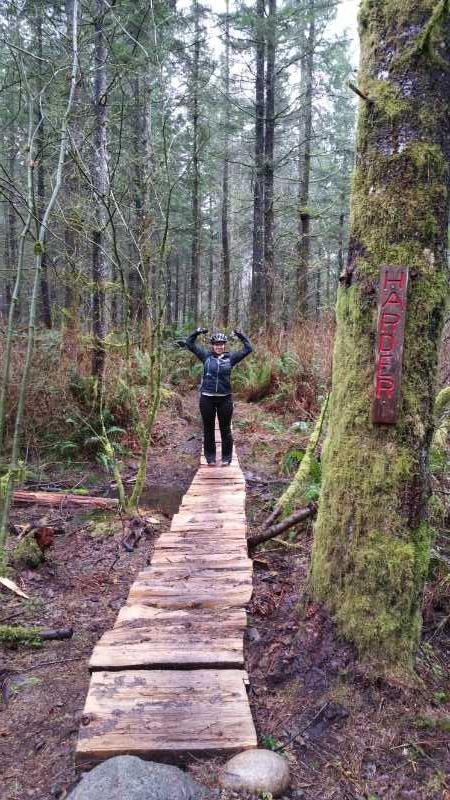 A person stands triumphantly on a wooden bridge in a forest, surrounded by tall trees and lush greenery. The individual is wearing a helmet and biking attire, striking a victory pose with arms raised. A sign on a nearby tree reads "HADDER." The scene captures the essence of outdoor adventure and exploration. Lake Sawyer mountain bike trail.