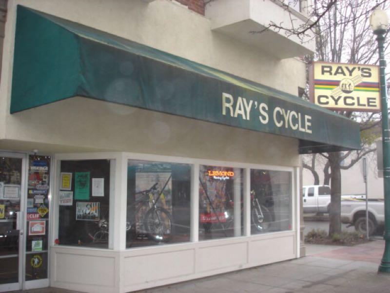 Storefront of Ray's Cycle, featuring a green awning with the name displayed prominently. The front windows showcase bicycles and various accessories, with signage visible on the door. A lamp post is nearby, and the street is lined with trees.